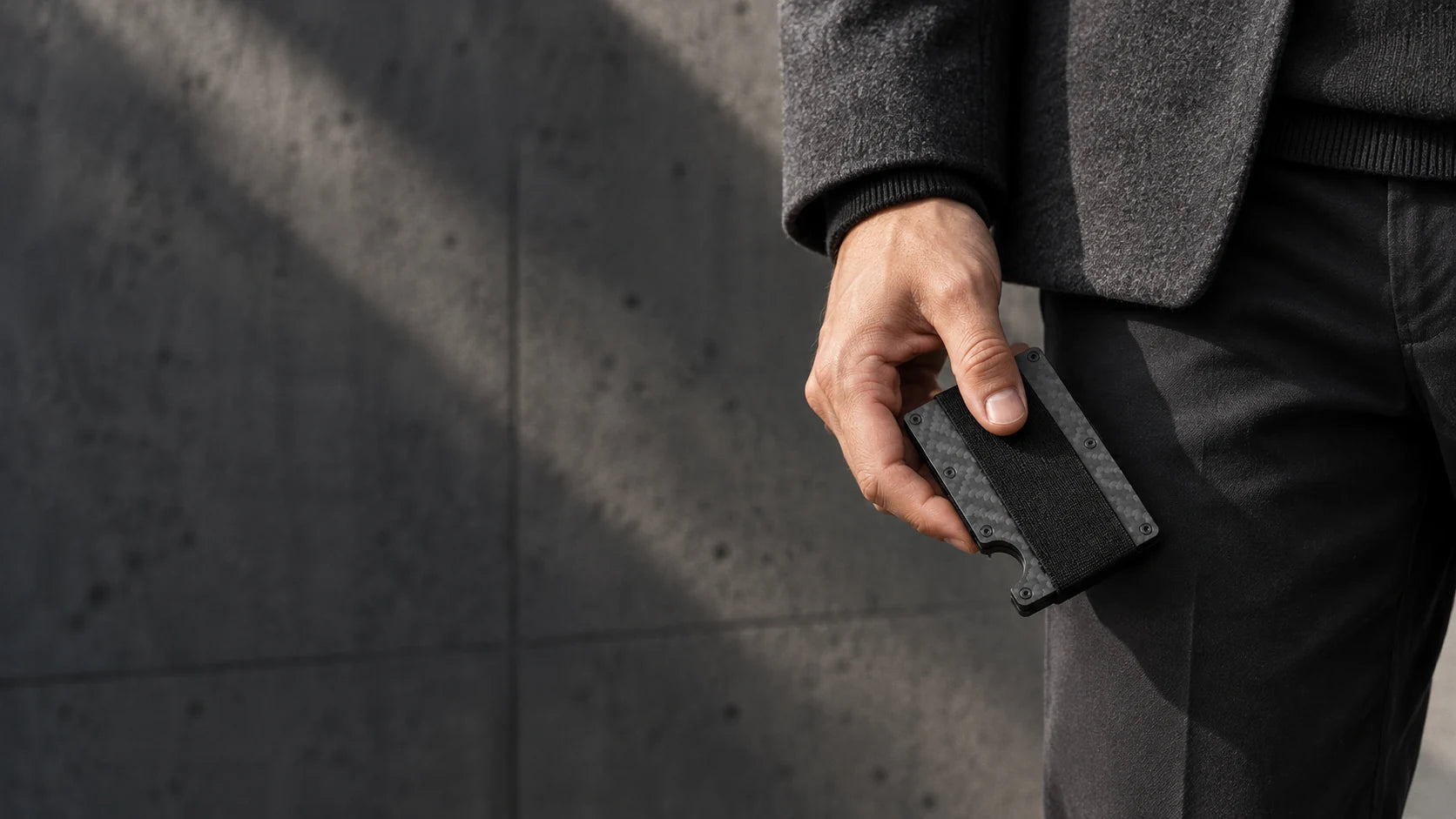 A compact carbon fiber wallet held beside a coat and trouser pocket